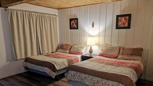 A room with two beds, wooden flooring, a window with curtains, and a nightstand with a lamp. - Cedars Motel (Princeton)