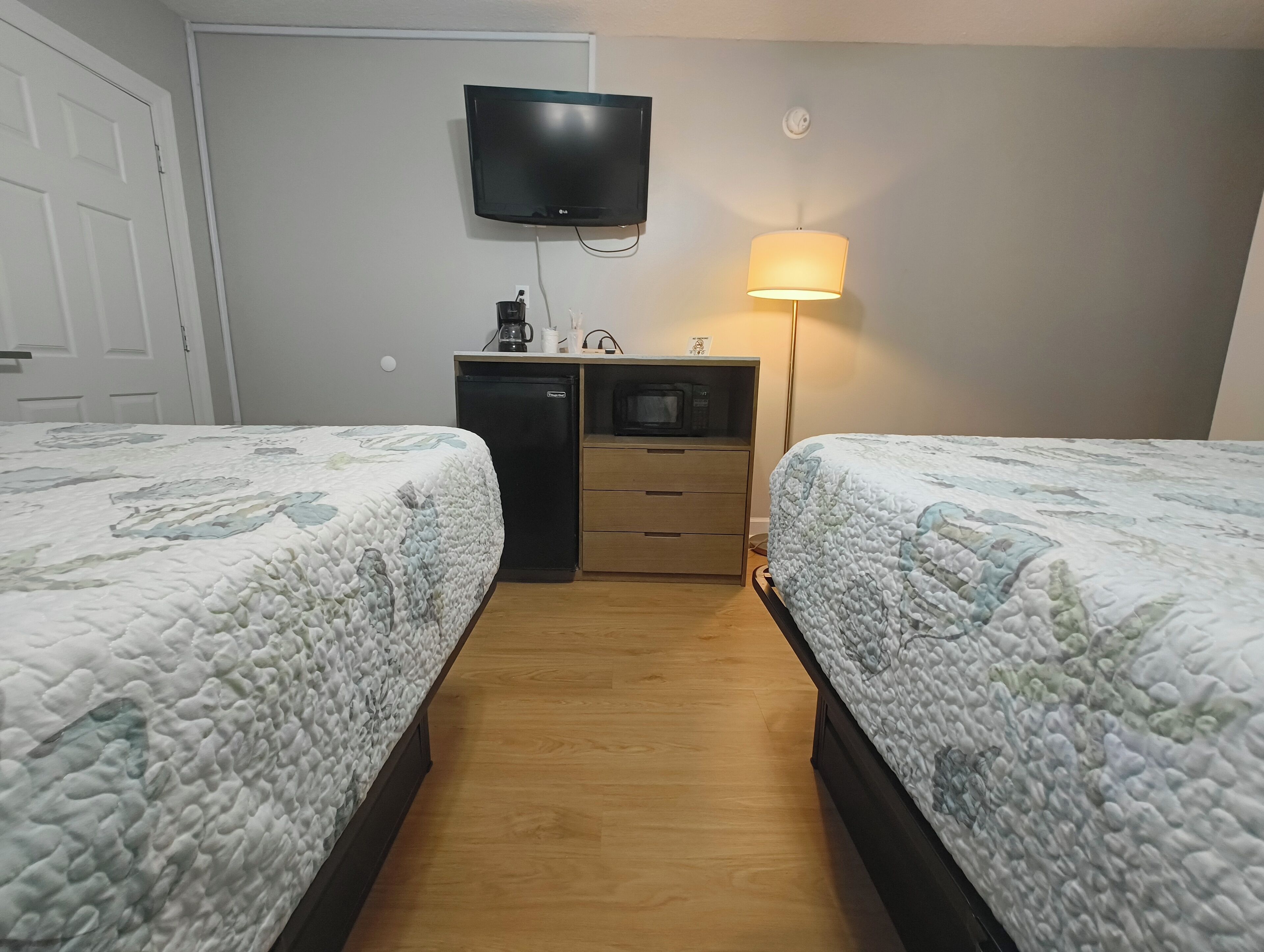 A hotel room with two beds, a TV mounted on the wall, a desk with a microwave, and a lamp.
