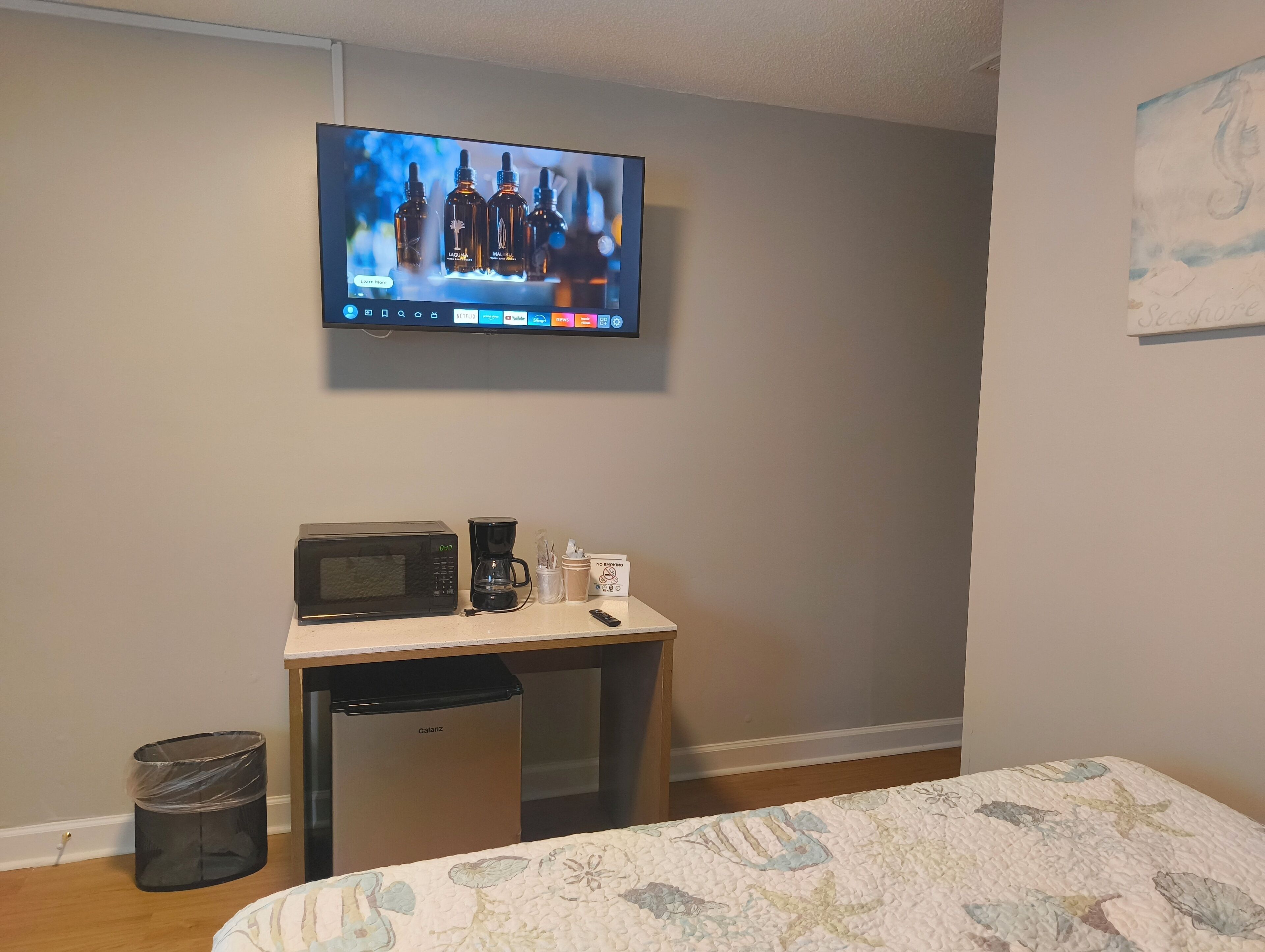 A hotel room with a television mounted on the wall, a small desk with a microwave, coffee maker, and a trash bin.