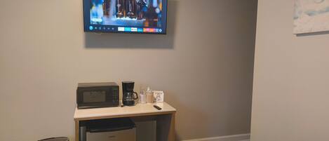 Habitación de hotel con televisión montada en la pared, un escritorio pequeño con microondas, cafetera y un cubo de basura.