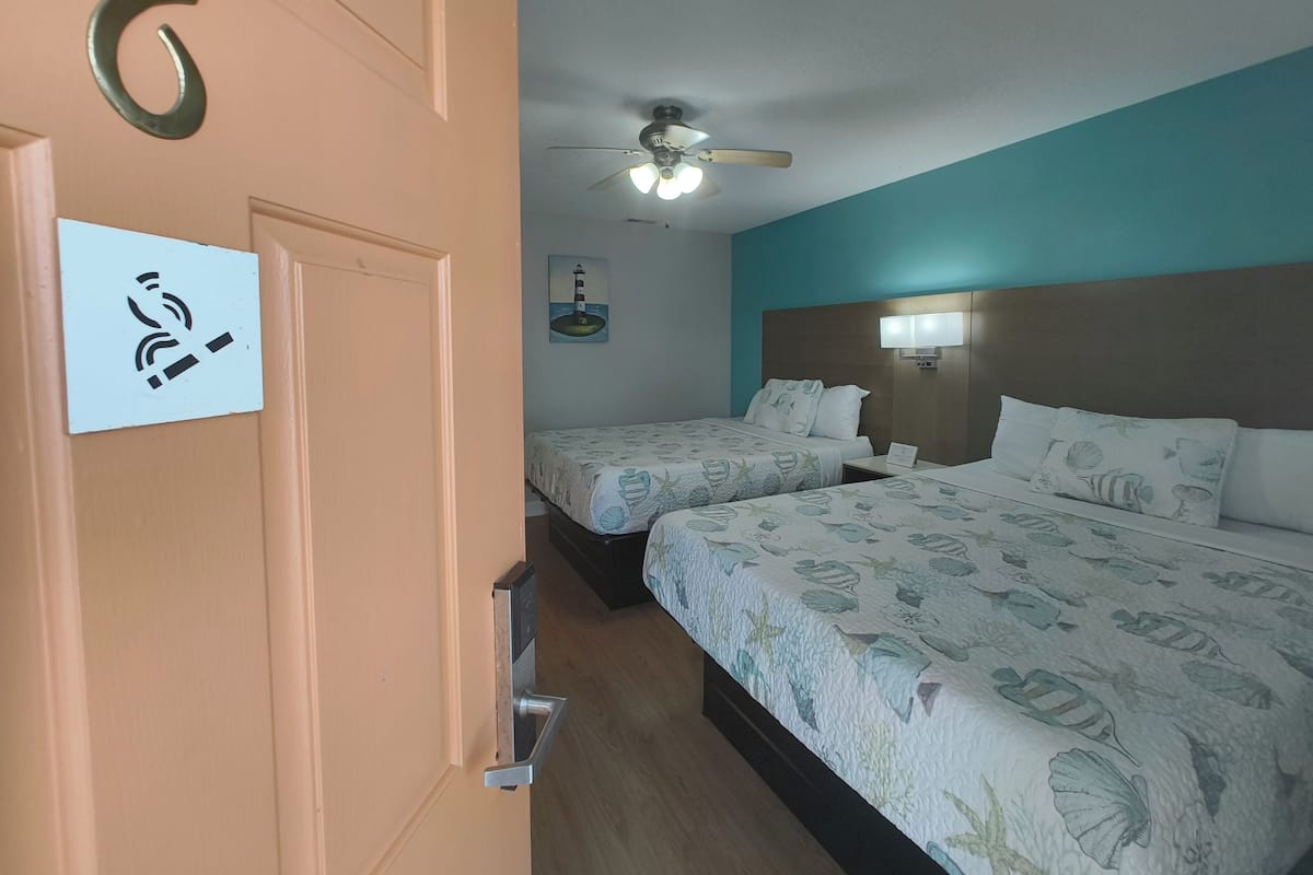 Habitación de hotel con dos camas, un ventilador de techo, un cuadro de un faro y un letrero que indica una zona para fumadores.