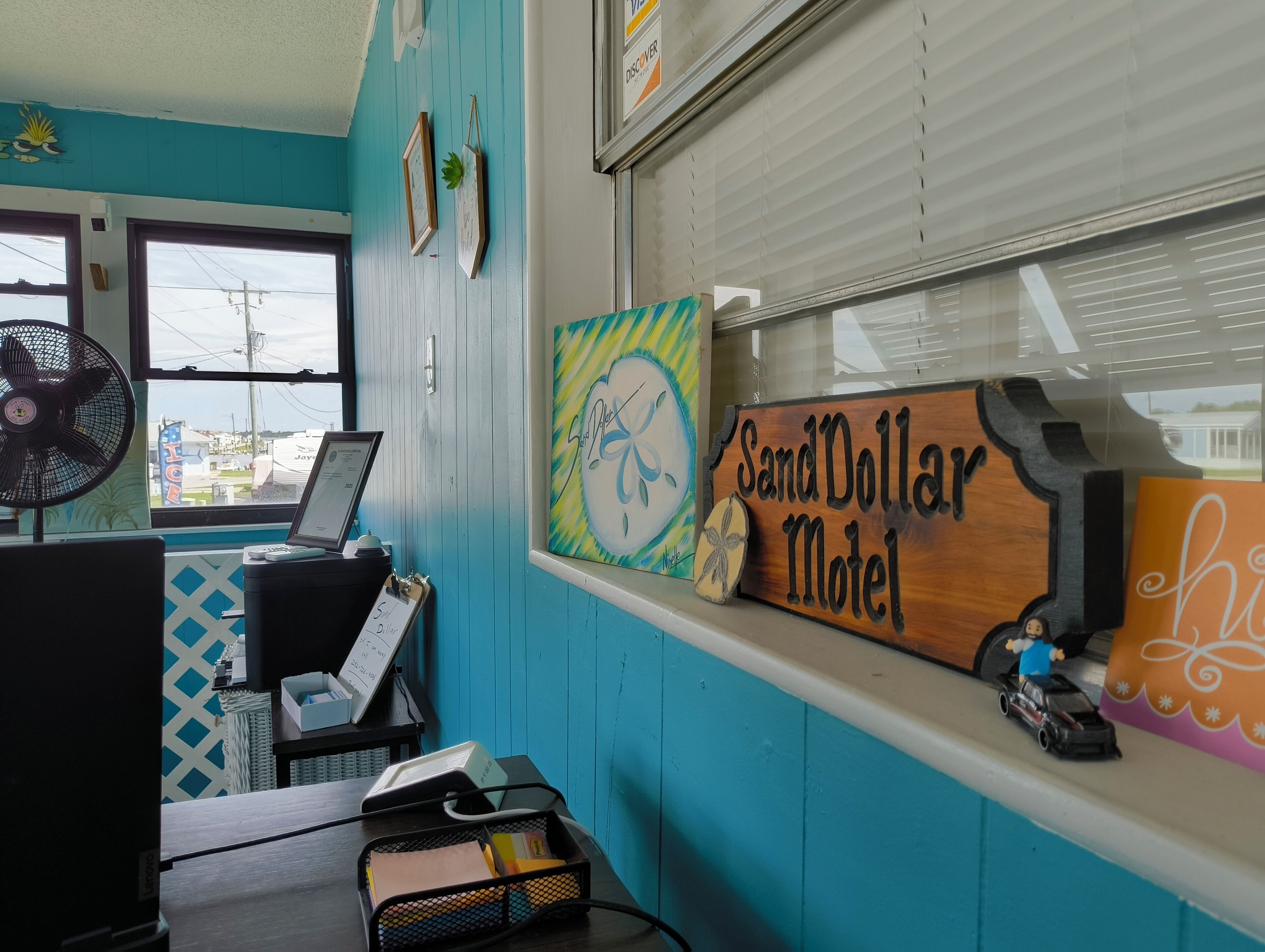 A room with a desk, a computer, a fan, and a window with a view of buildings. There are two framed pictures on the wall and a sign that reads "SandDollar Motel".