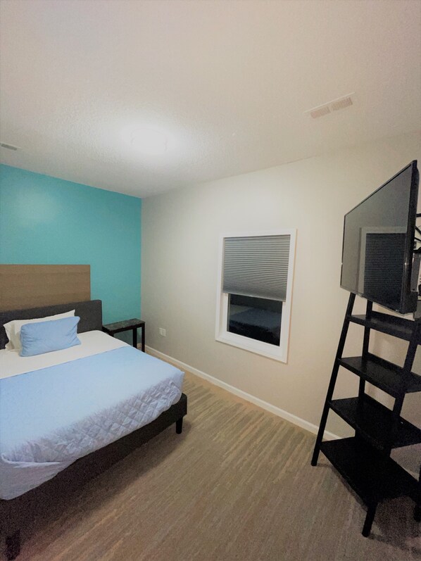 A bedroom with a bed, a TV on a stand, a window with blinds, and a small table.