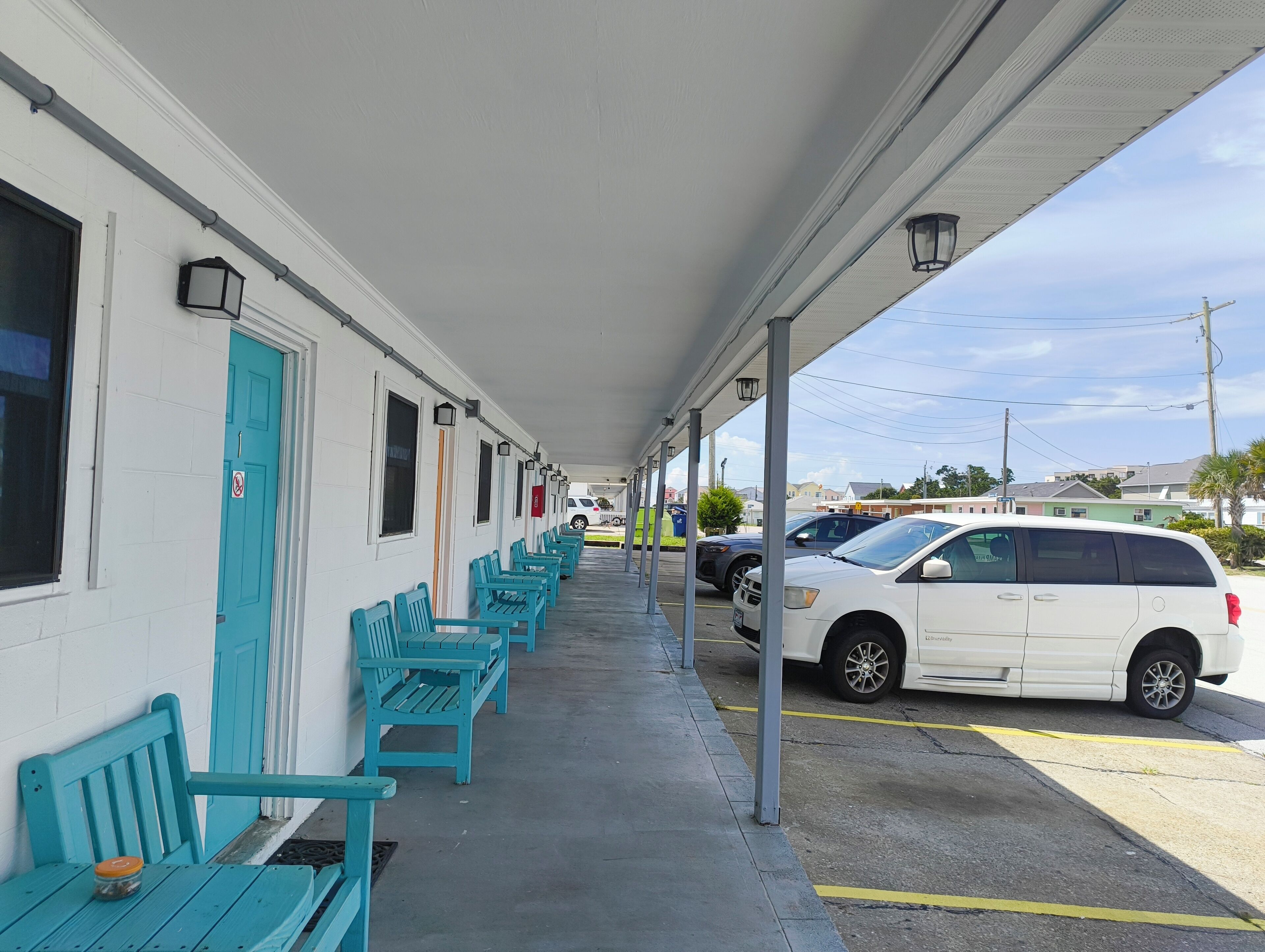 A motel with a covered porch, rows of turquoise chairs, and a parking area with cars.