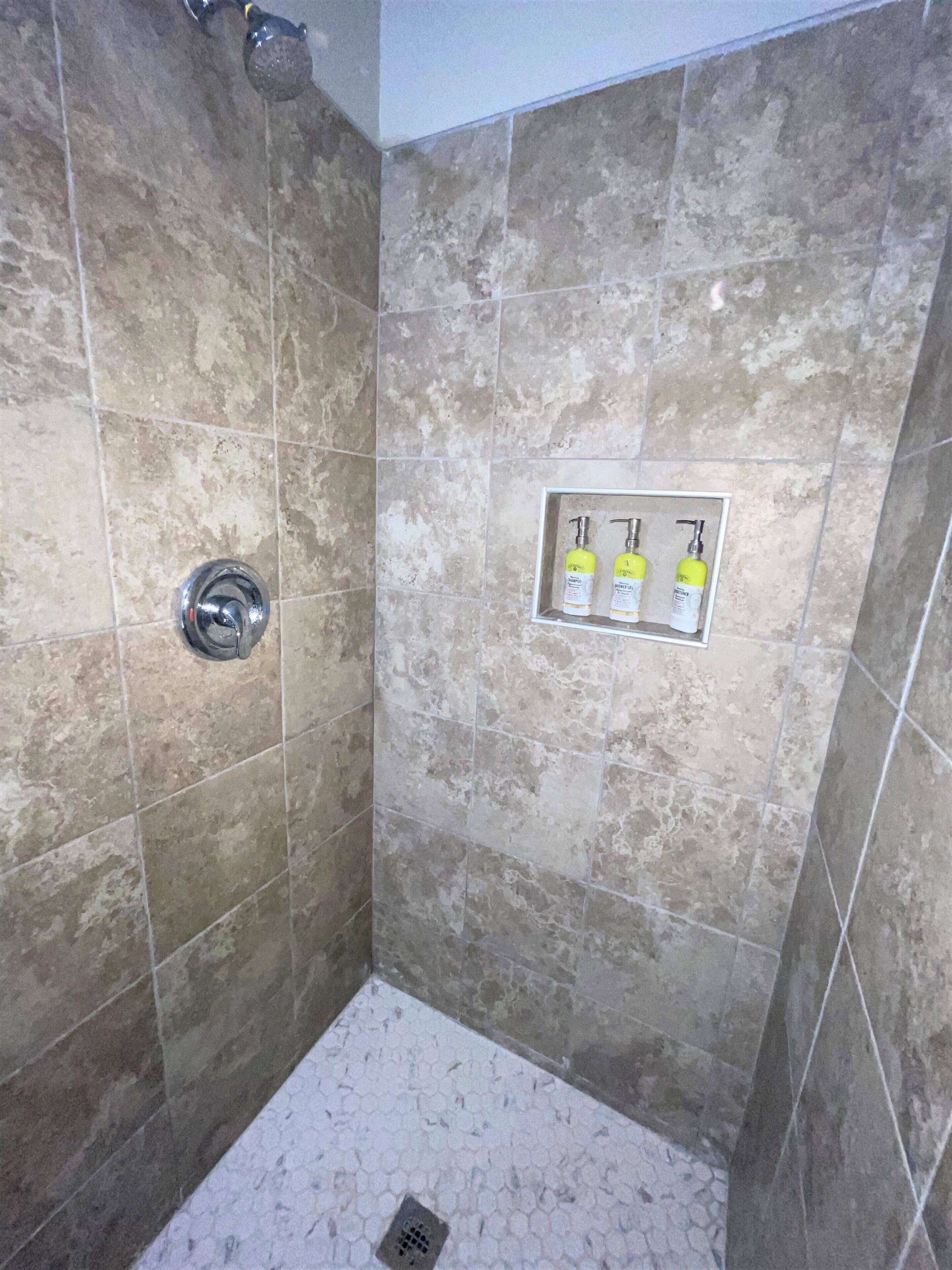 Une douche aux carreaux beiges, un pommeau de douche chromé et une étagère avec trois bouteilles.