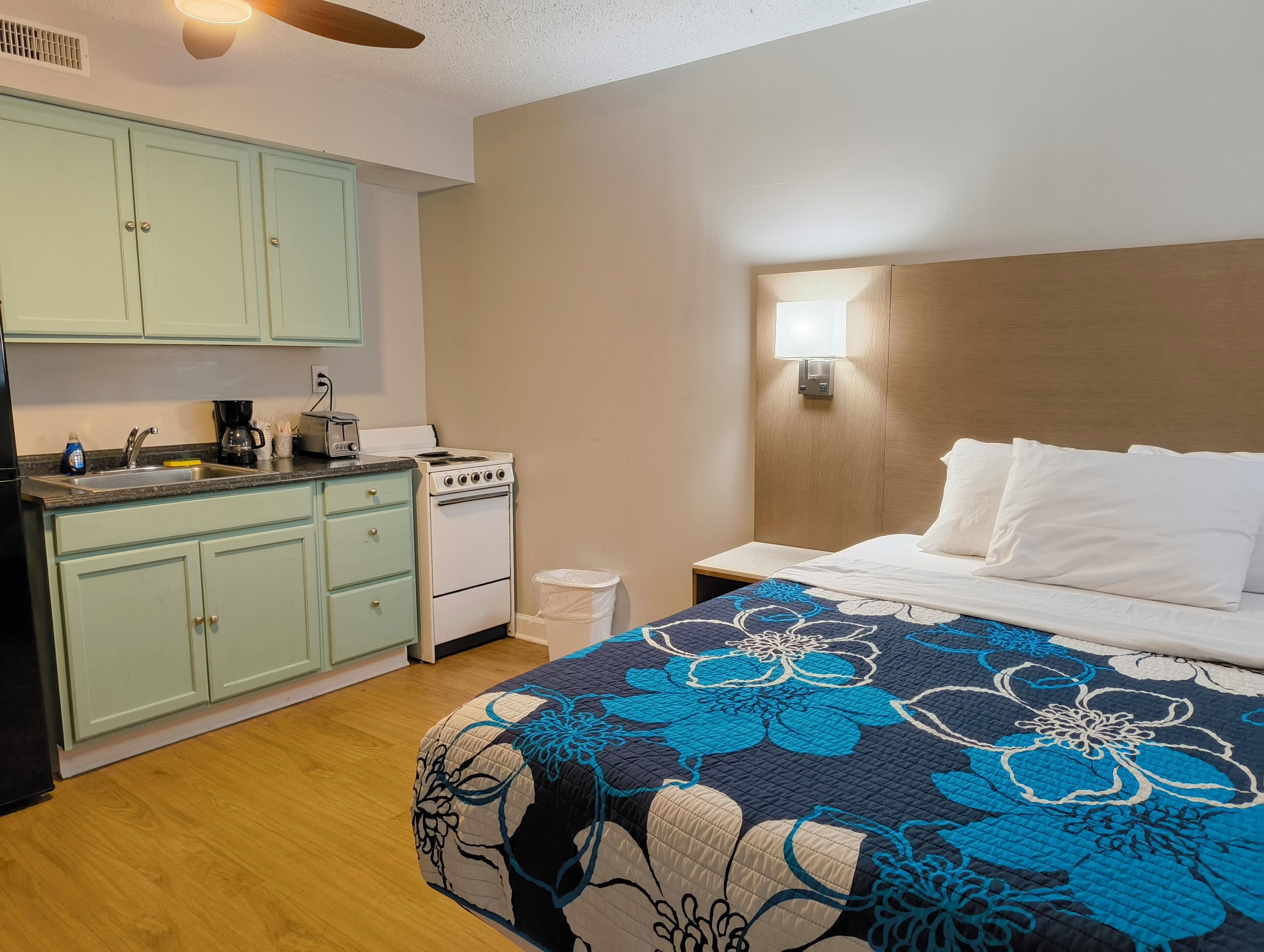 A compact hotel room with a kitchenette, a bed with a floral-patterned duvet, and a nightstand with a lamp.