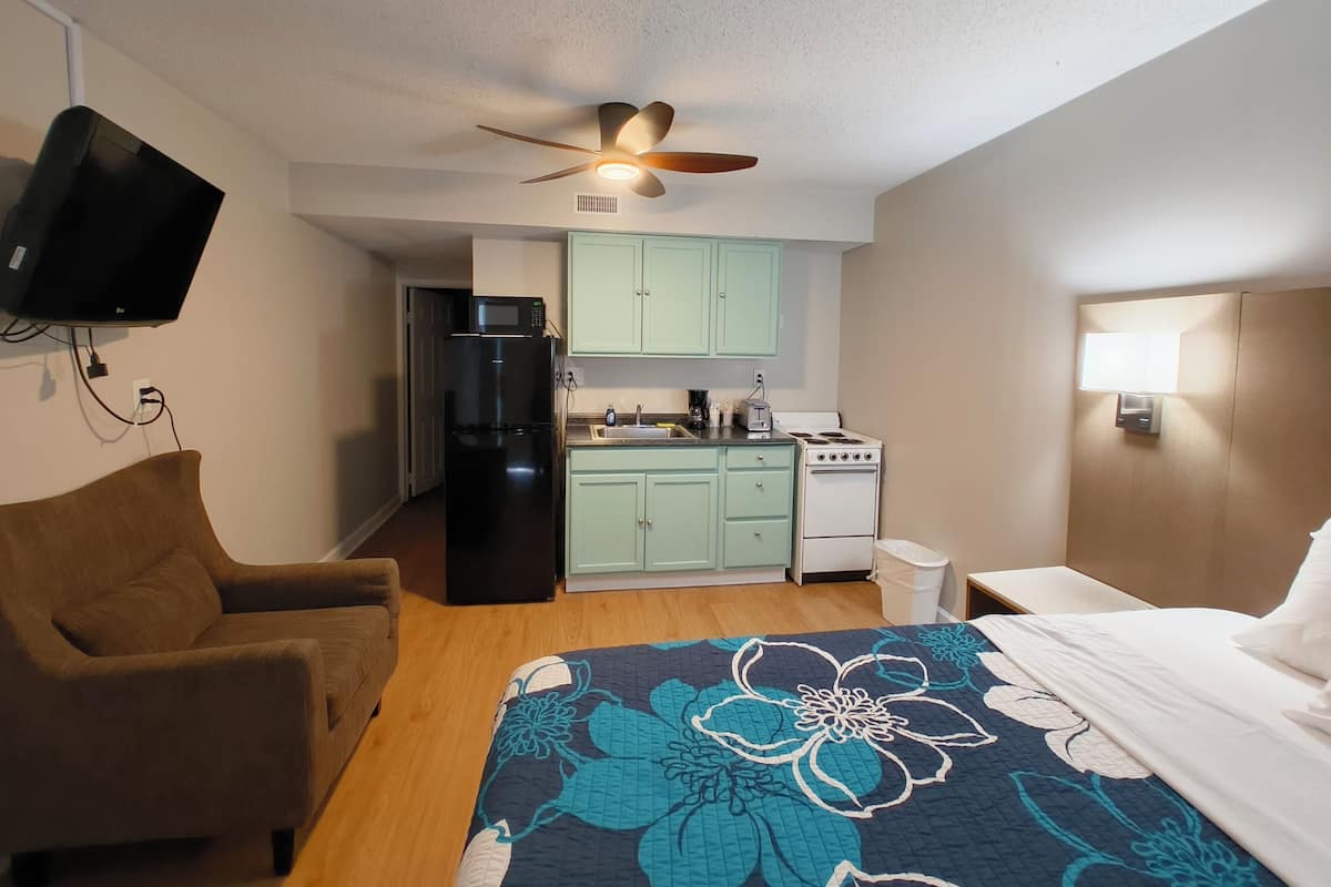 Habitación de hotel compacta con cama, sofá, kitchenette equipada con refrigerador, estufa y microondas, ventilador de techo y televisor montado en la pared.