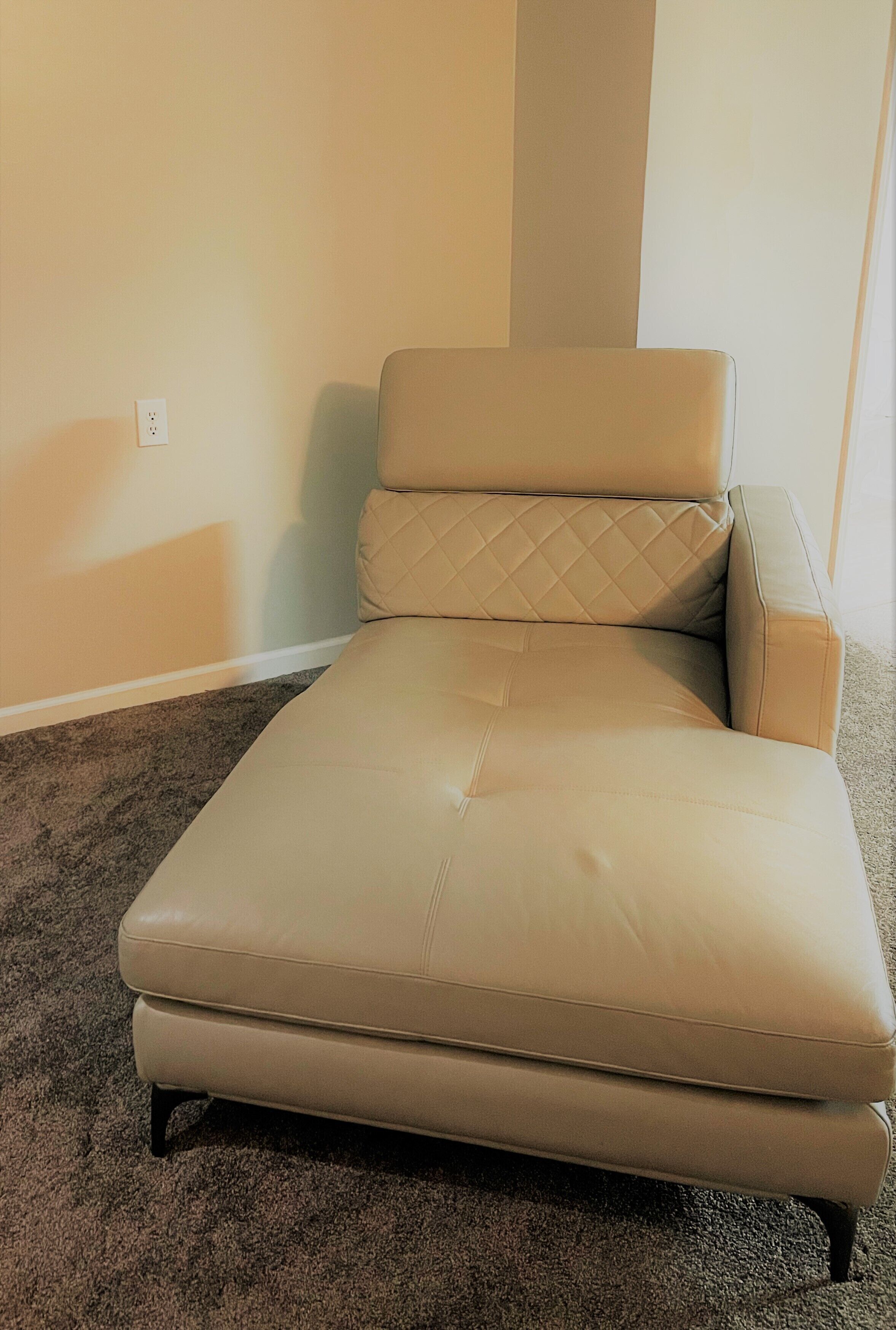 A single beige leather sofa with a quilted backrest and armrests, positioned against a light-colored wall with a power outlet visible.