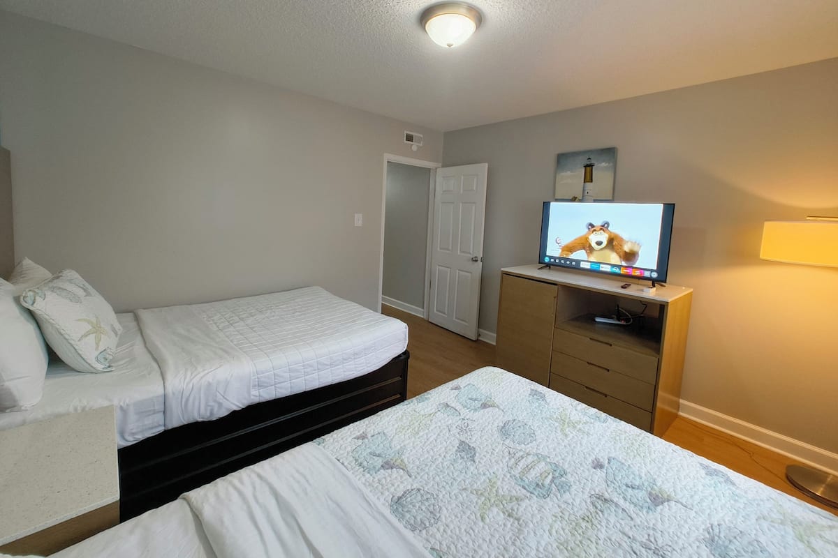 Un dormitorio con dos camas, un televisor sobre un aparador de madera y una puerta que da a otra habitación.