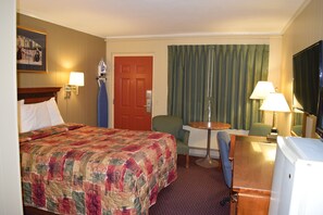 A hotel room with a large bed, a desk with a lamp, a chair, a television, and a door.