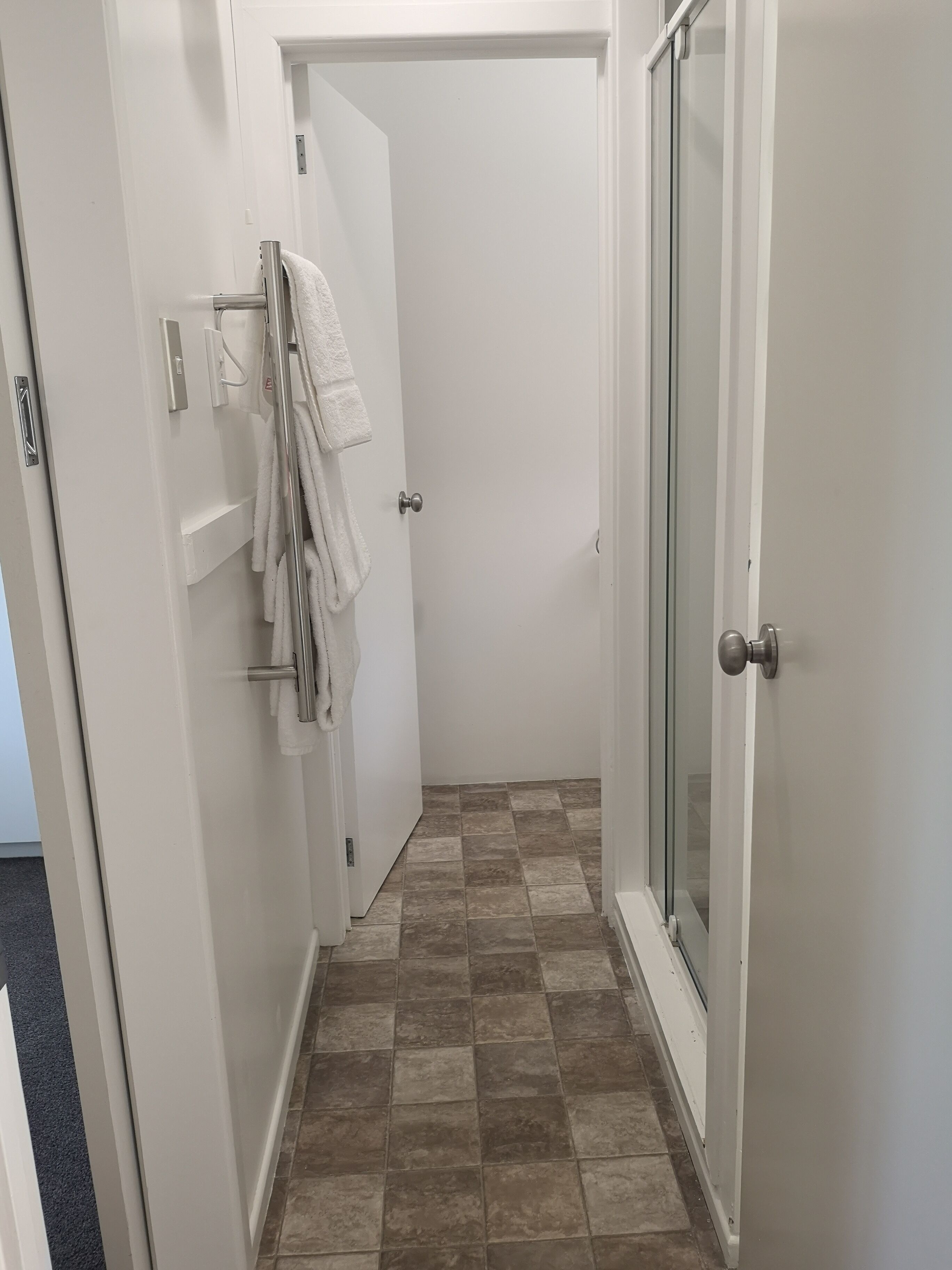 A narrow hallway with a tiled floor, a white door with a towel hanging on the door handle, and a glass shower door.