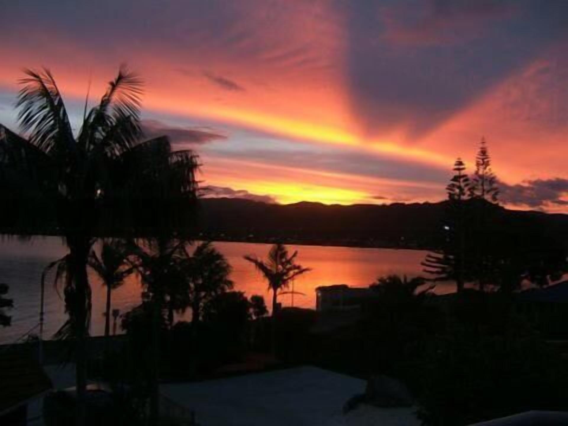 Una vista al atardecer sobre un cuerpo de agua, con palmeras y una cadena montañosa distante.