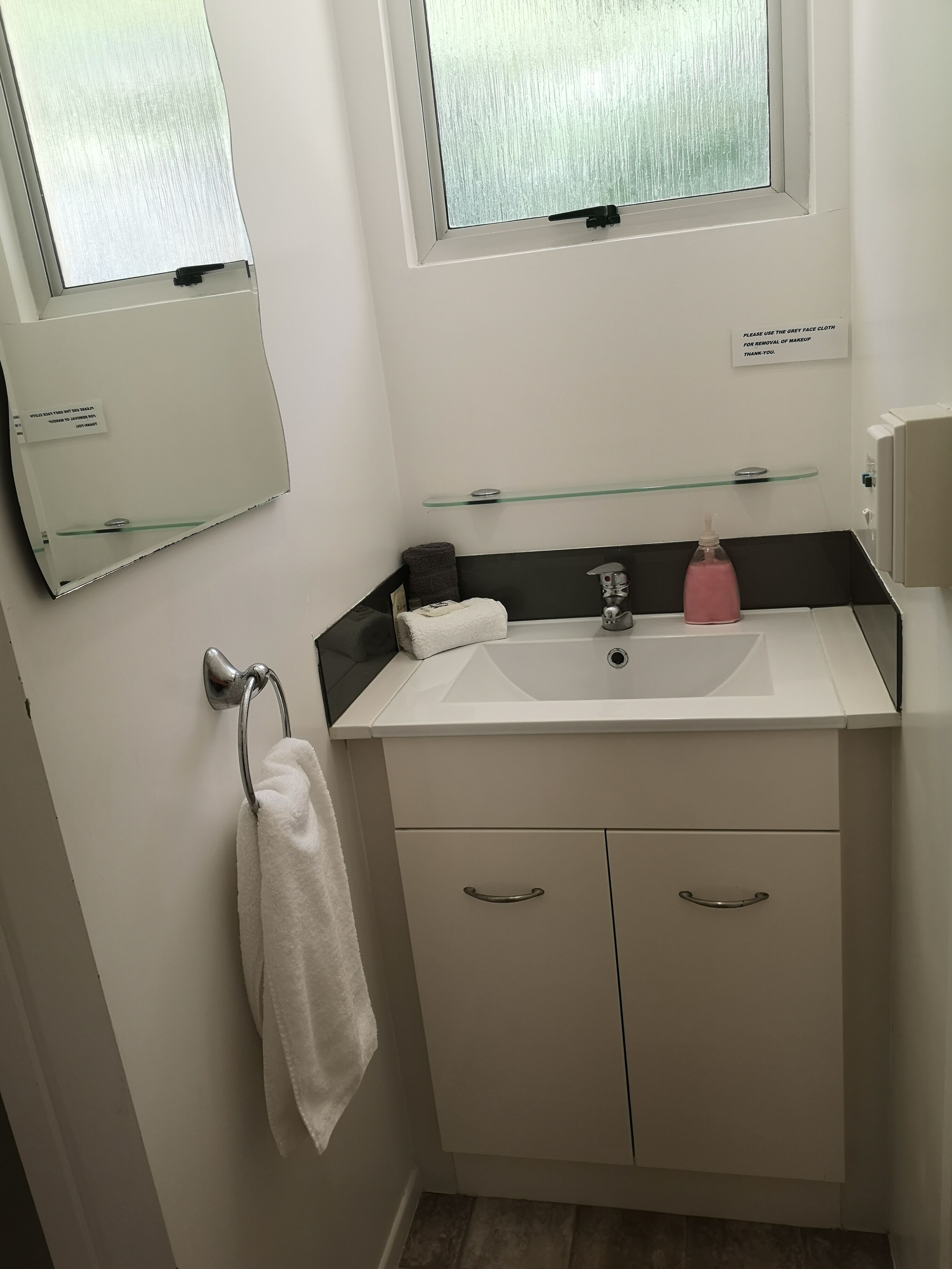 A bathroom with a white sink, a glass shelf, a towel rack, and a sign instructing to use grey face cloth for makeup removal.