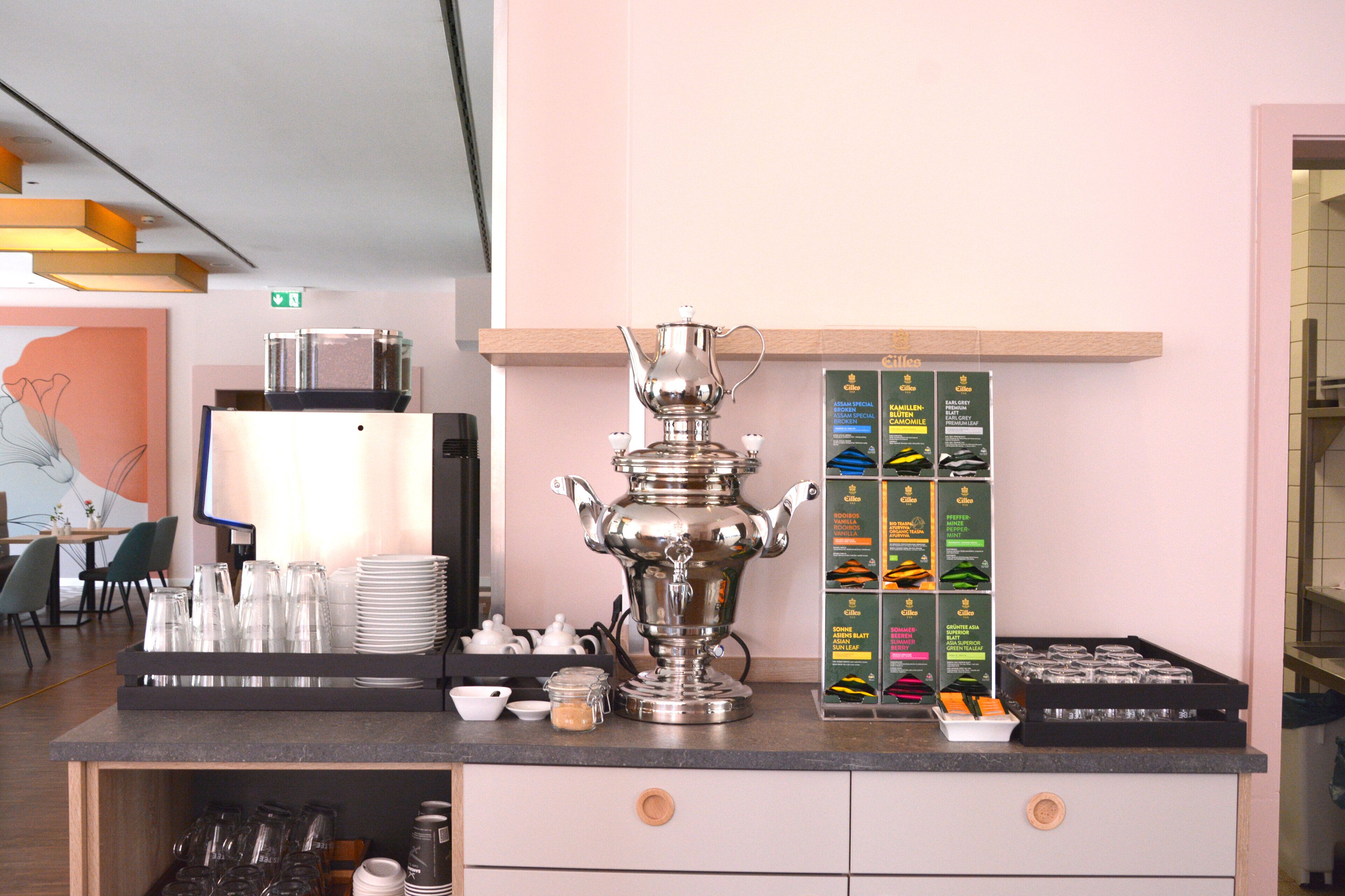 A kitchen counter with a large silver teapot, stacks of white plates, and a display of packaged food items.