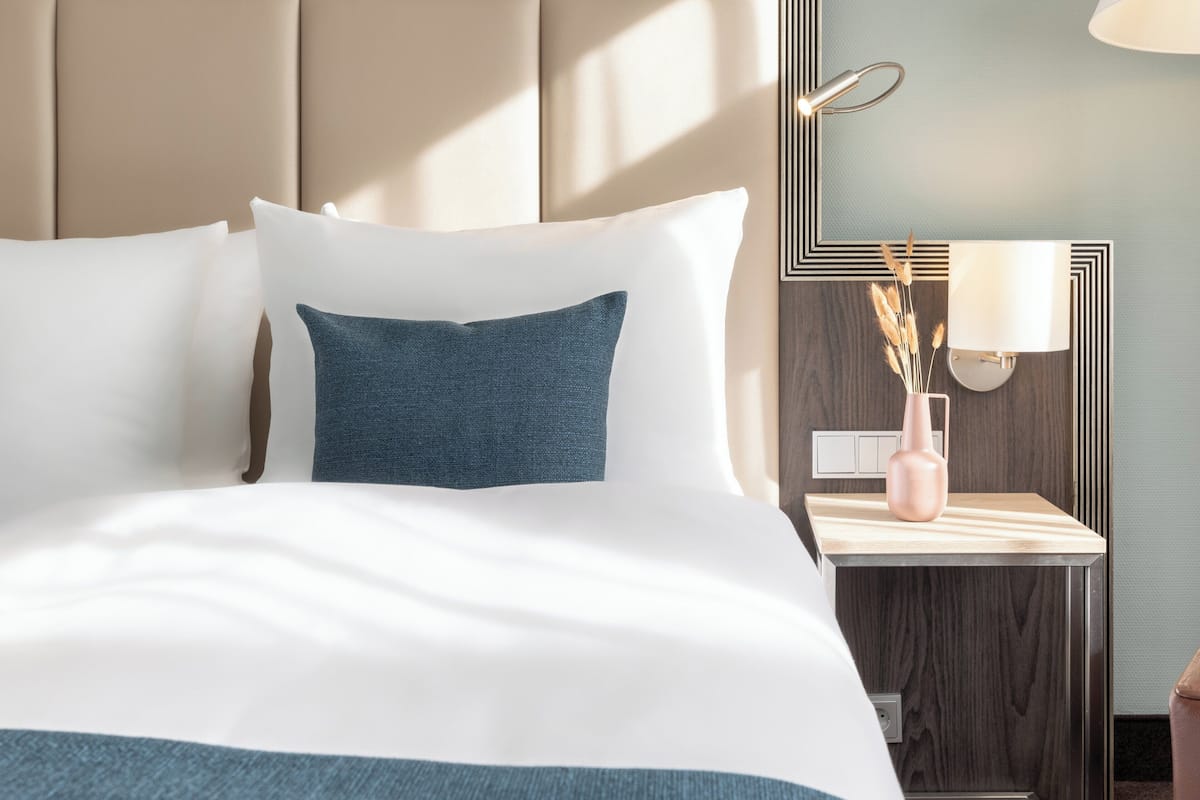 Een hotelkamer met een bed, nachtkastje, lamp en een vaas met gedroogde planten.