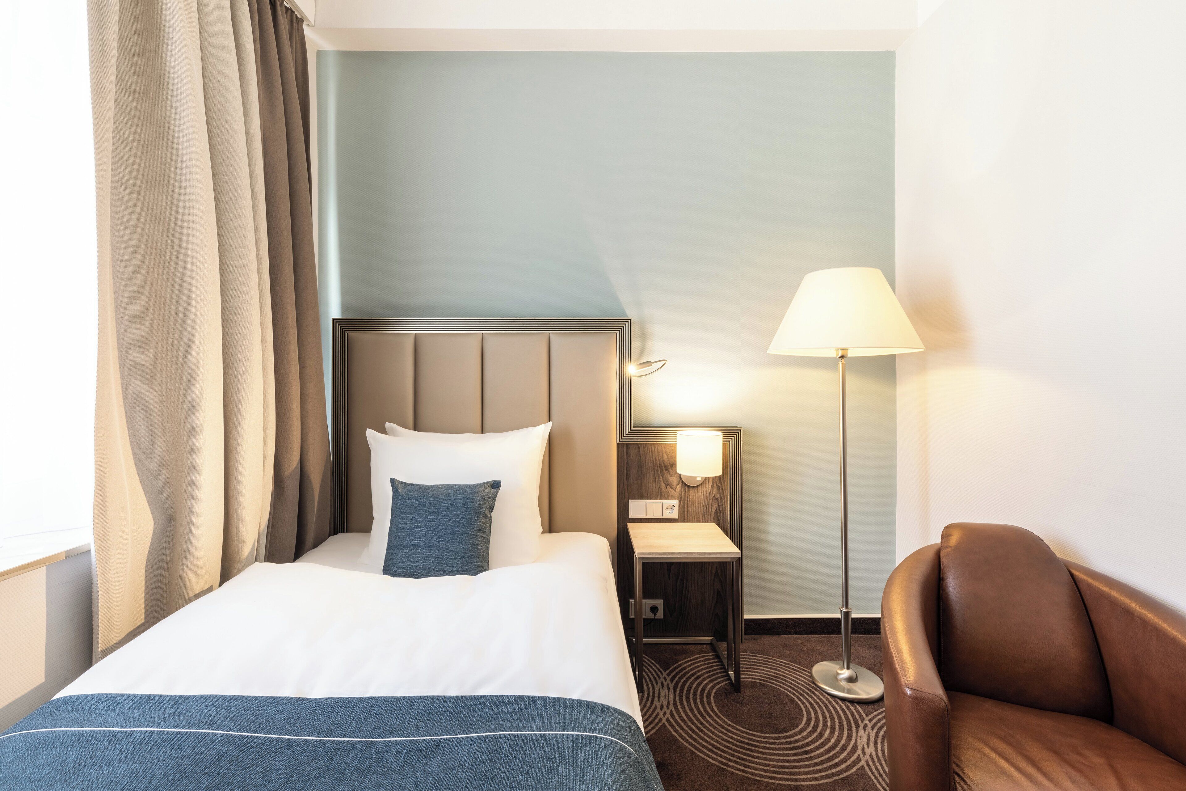 A hotel room with a bed, a nightstand, a lamp, a chair, and a patterned rug.