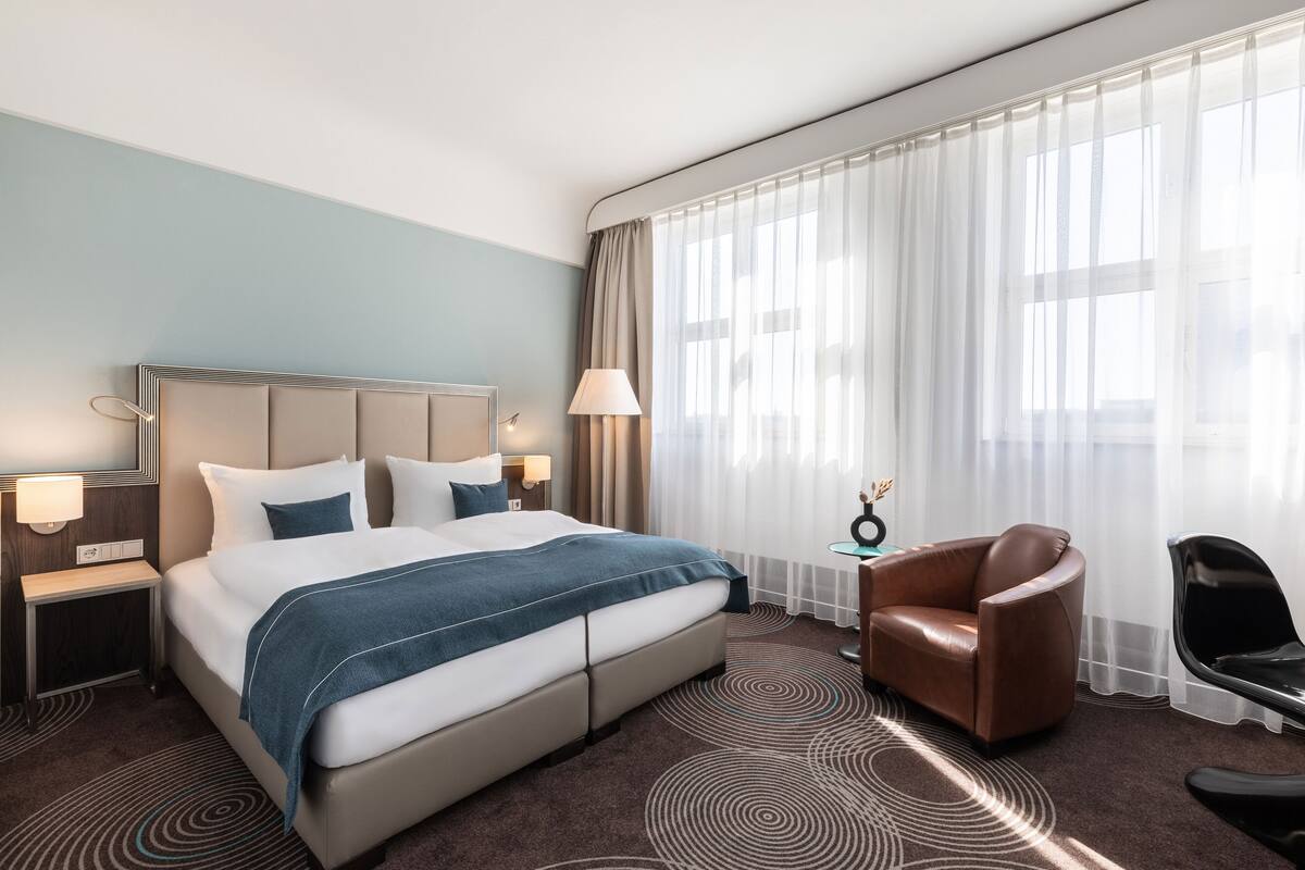 Een hotelkamer met een groot bed, een stoel, een bureau en een raam met dunne gordijnen.