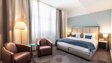 A hotel room with a large bed, two chairs, a small table, and a nightstand with lamps.