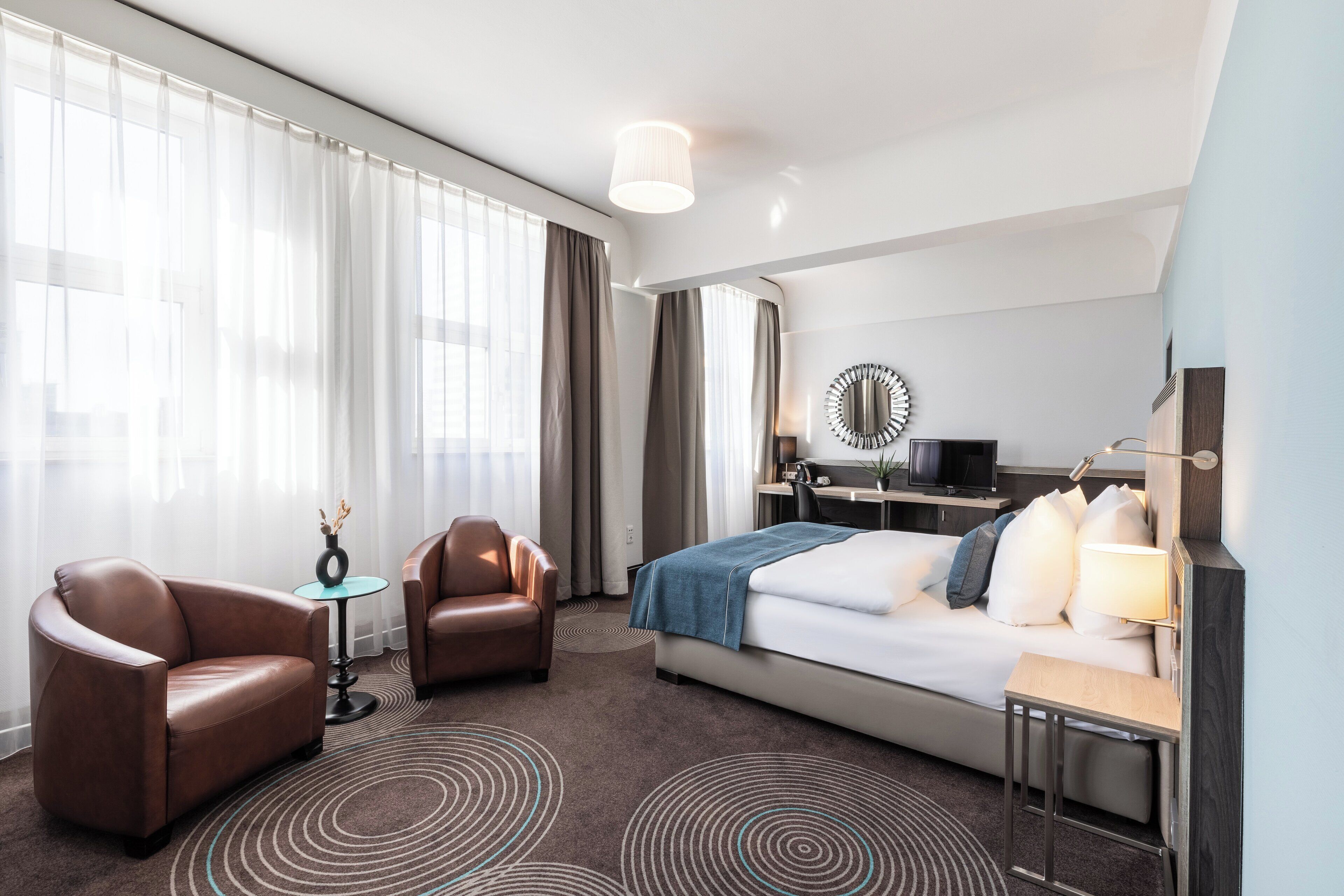 Une chambre d’hôtel comprenant un lit, deux fauteuils en cuir, un bureau avec un ordinateur, un miroir et une petite table avec une lampe.