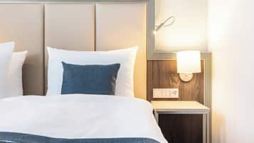 Un letto rifatto con cuscini bianchi e blu, un comodino in legno con una lampada a parete e una presa elettrica.