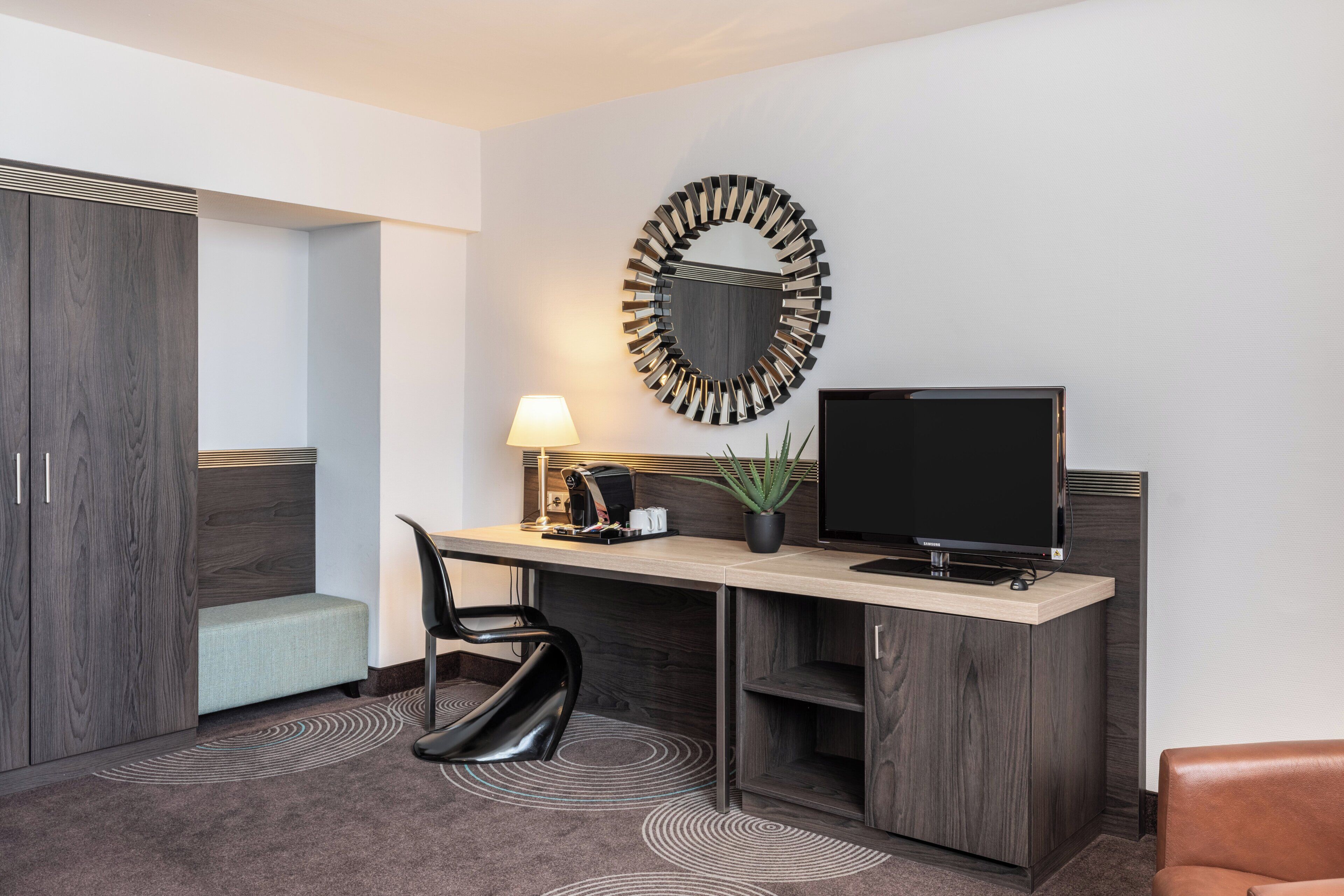 A modern hotel room with a desk, chair, TV, and a decorative mirror.