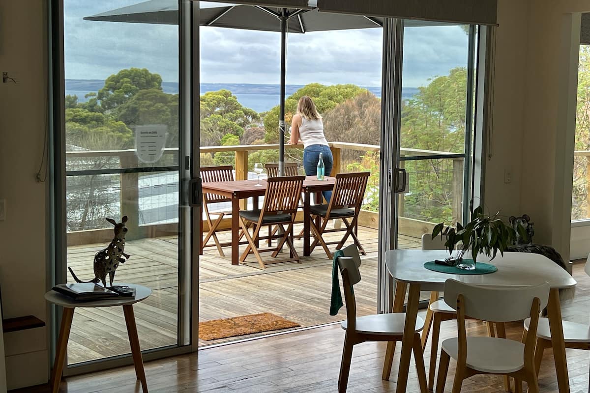 A spacious room with a large sliding glass door leading to a balcony with a wooden deck, a dining area with a table and chairs, and a small table with a decorative animal figure.
