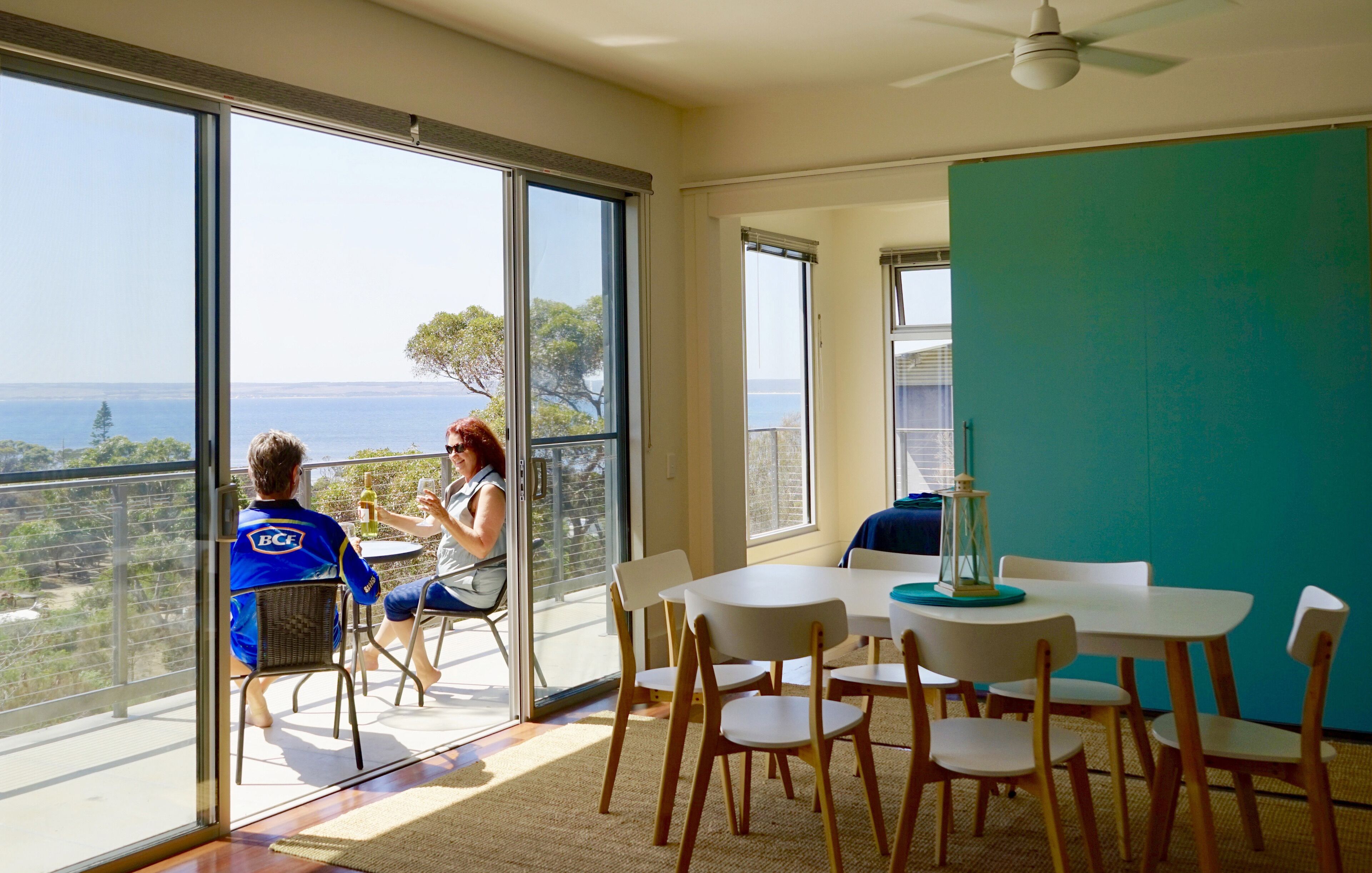 Un balcon avec une table et des chaises, une personne portant une chemise bleue, et une vue sur la mer.