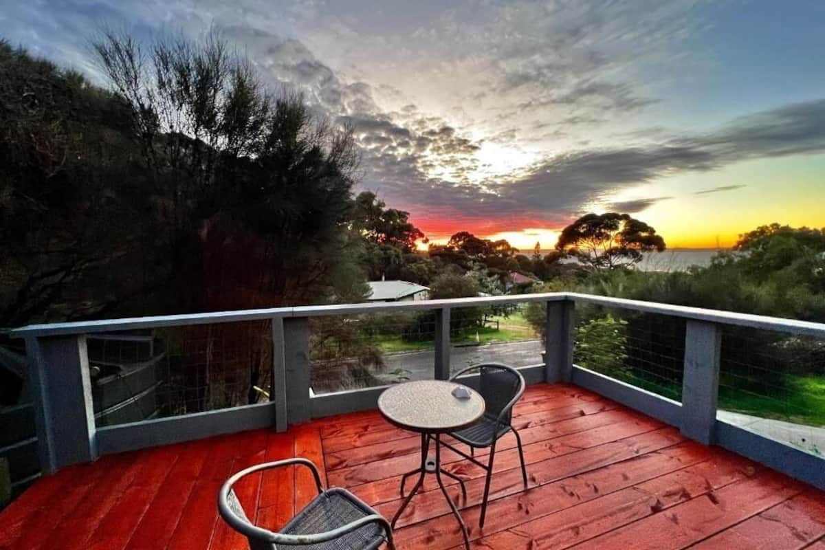 A wooden deck with two chairs and a table, overlooking a sunset with trees and a road in the background.