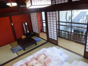 Una habitación japonesa tradicional con suelo de tatami, un futón, una mesa de madera con un espejo y puertas correderas que dan a un balcón con vistas a un edificio tradicional.