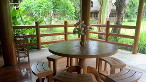 A wooden round table with benches, a vase with flowers, and a view of greenery and a wooden building through a bamboo fence. - Saksuay Klangdoi Resort (Mae Hong Son)