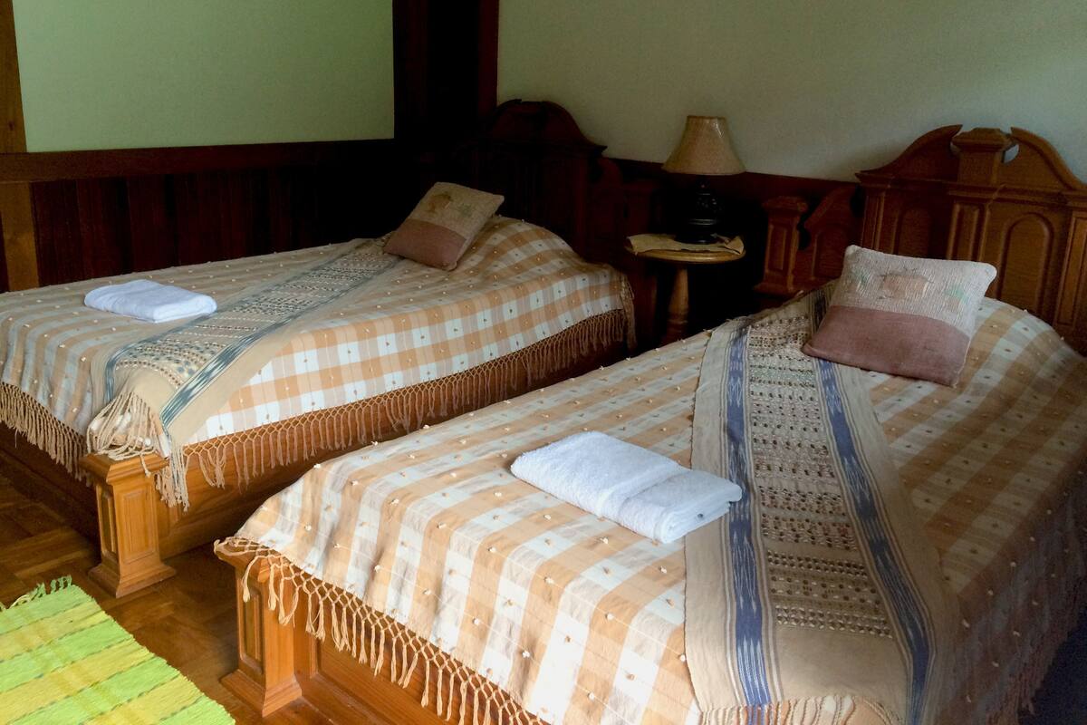 Two single beds with checkered bedding and pillows, a wooden headboard, a small table with a lamp, and a green striped rug.