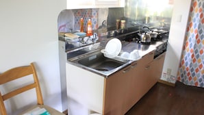 A compact kitchen with a sink, stove, and cabinets. A chair and a table with a paper are visible. - Tocotoco House (Nakafurano)