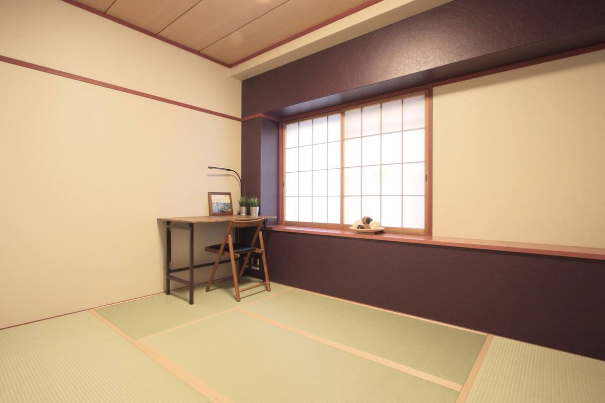 A tatami mat room with a low table and chair, a window with shoji screens, and a small shelf with items.