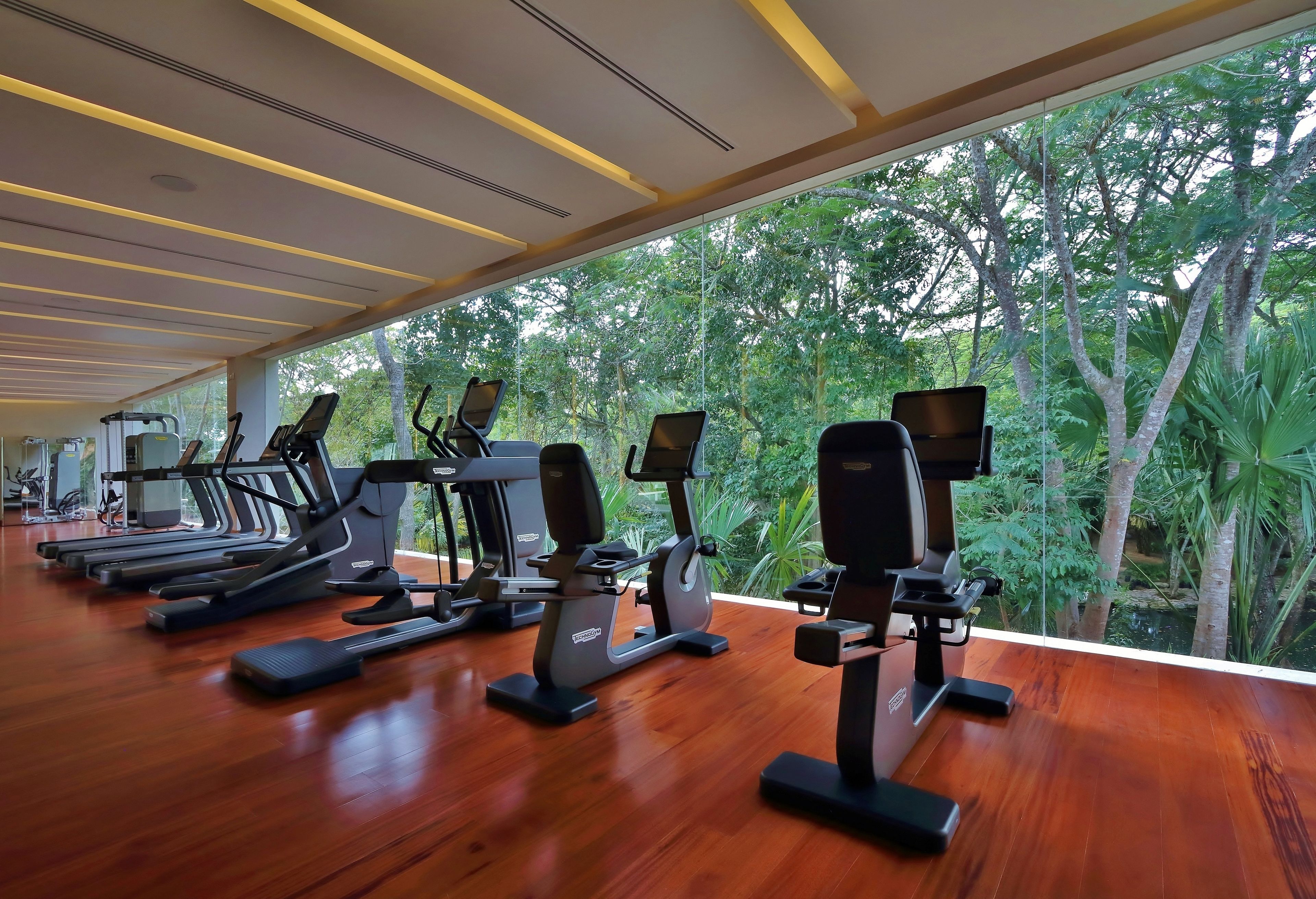 Une salle de sport équipée de tapis de course, de vélos elliptiques et de postes de musculation, avec une vue sur une végétation luxuriante grâce à de grandes fenêtres.