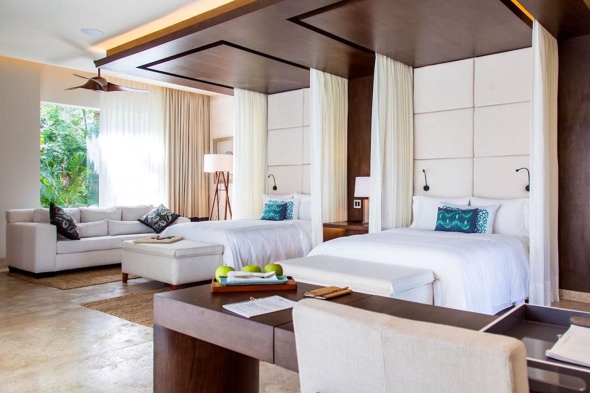 A hotel room with a bed, a sofa, a desk, and a view of greenery outside.