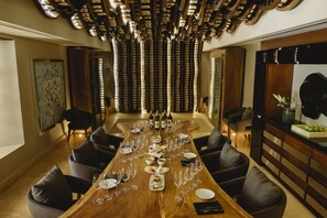 A long dining table set with wine glasses, plates, and cutlery, surrounded by upholstered chairs in a modern interior with a patterned ceiling and a large mirror on the right.