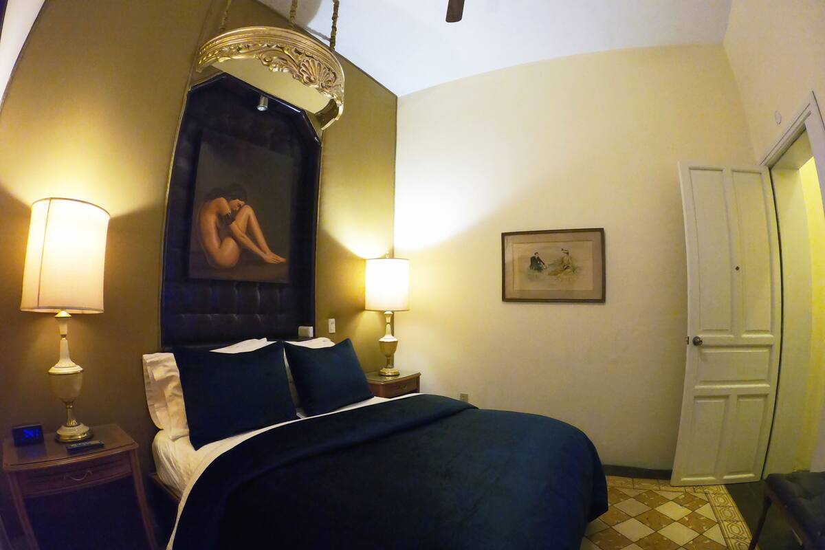 A hotel room with a bed, two bedside lamps, a nightstand, a framed picture, and a door.