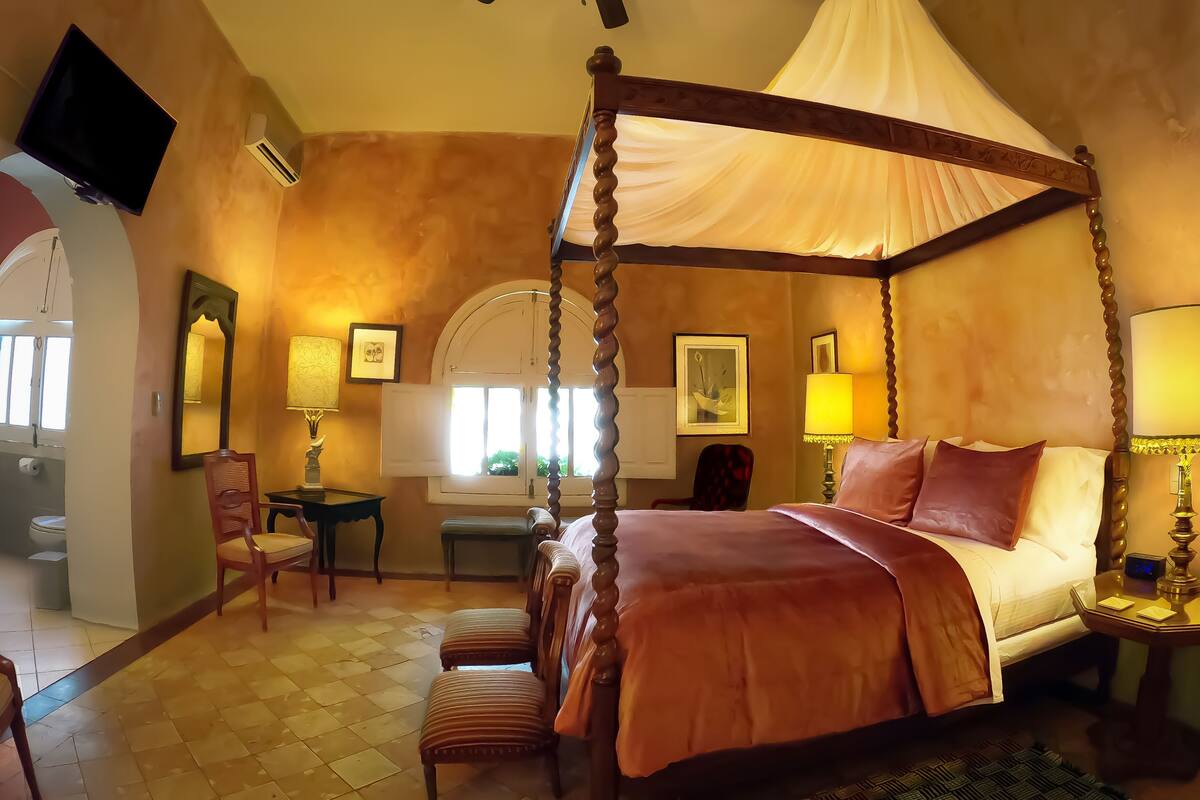 A four-poster canopy bed with a bench at the foot, a small table with a lamp, and a mirror on the wall.