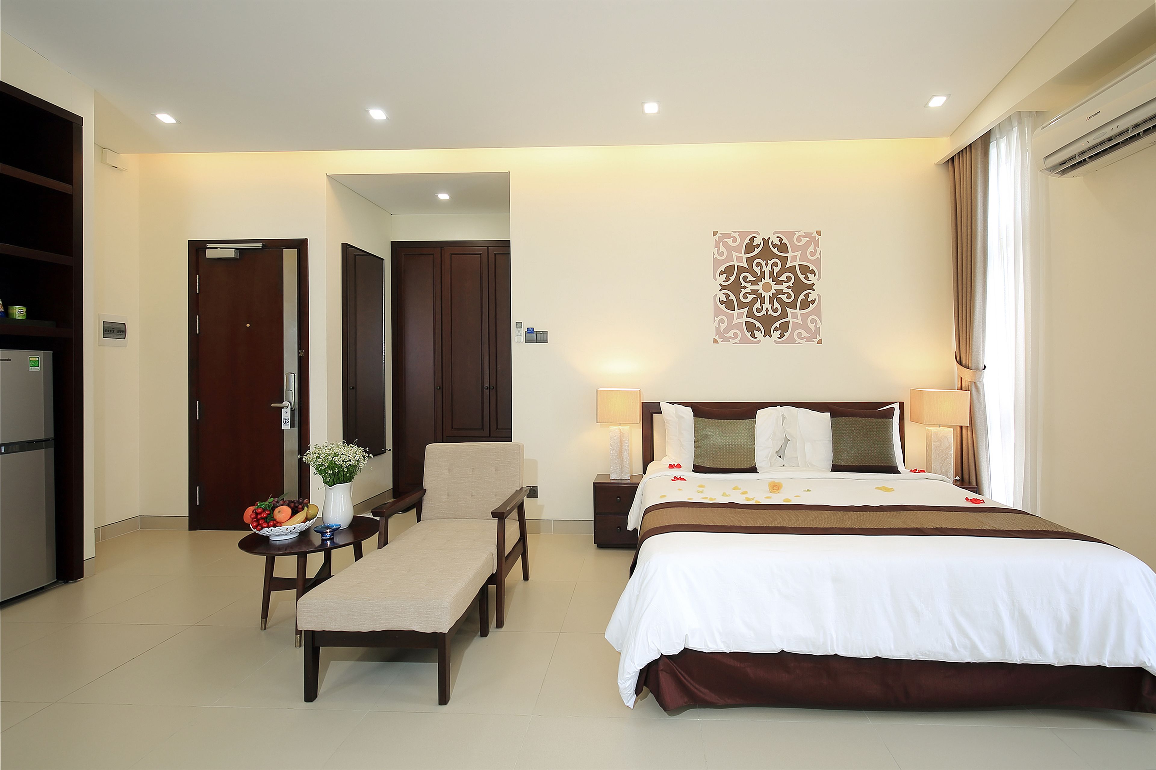Habitación de hotel con cama, mesitas de noche, un escritorio con silla, una pequeña mesa con un jarrón de flores y un refrigerador.
