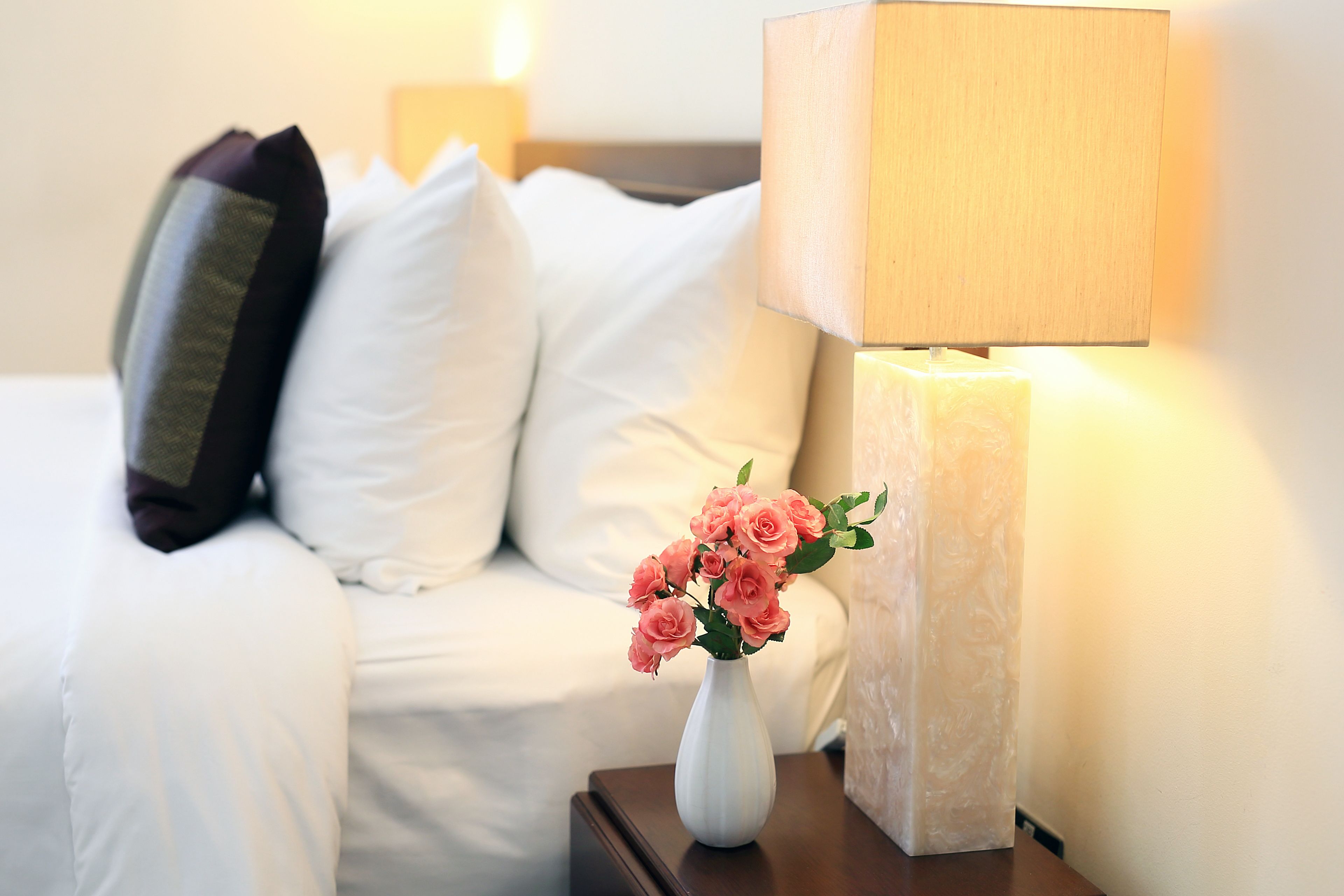 Un lit bien fait, avec du linge de lit blanc, un vase blanc contenant des fleurs roses, une table de chevet en bois et une lampe.