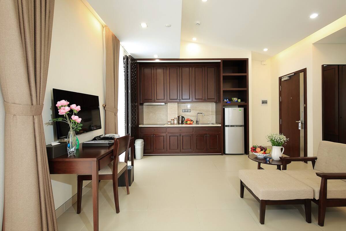 A modern kitchen with dark wooden cabinets, a built-in oven, and a refrigerator. There is a dining area with a wooden table and chairs, and a small seating area with a cushioned bench.