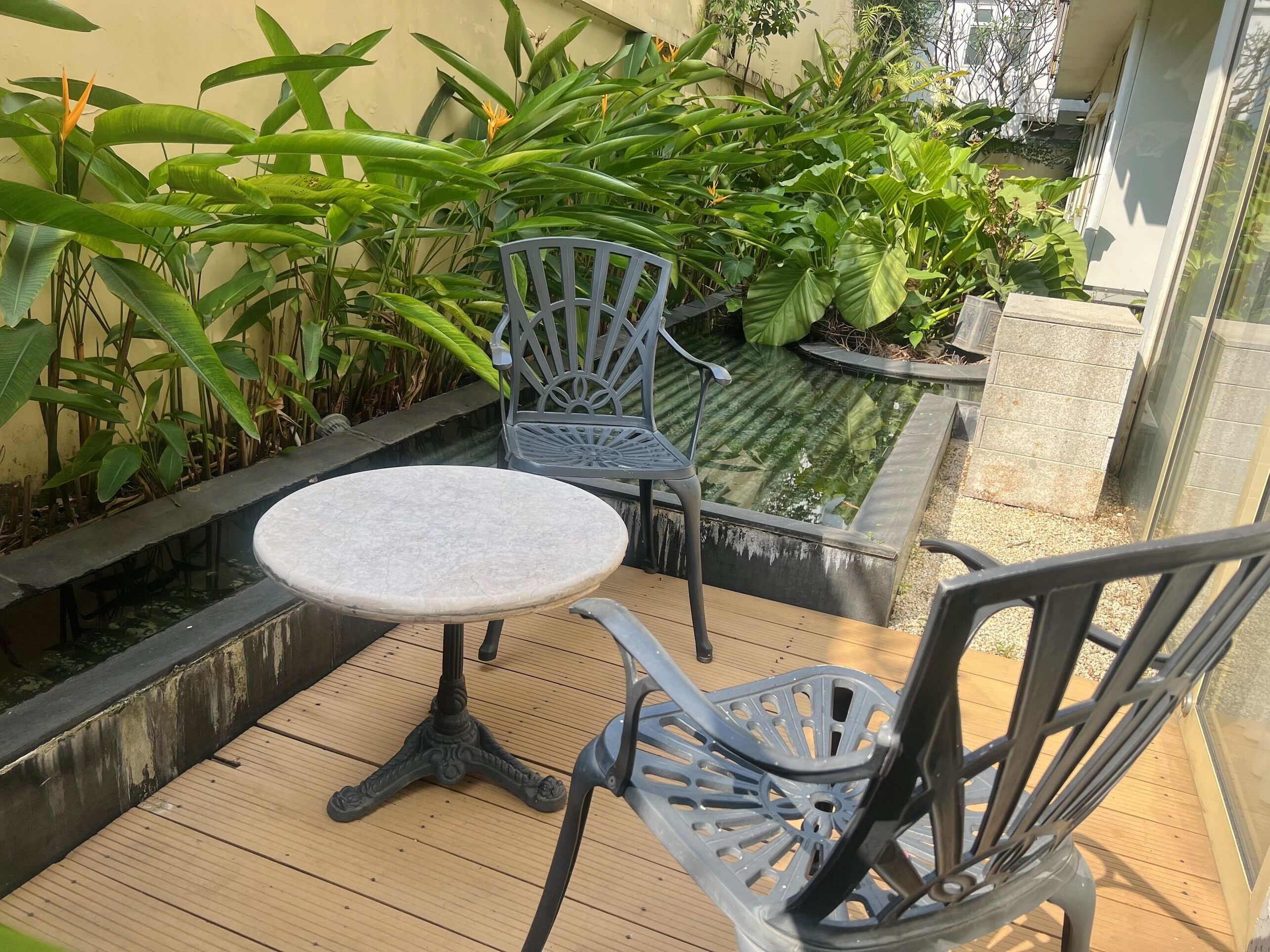 Una terraza de madera con una mesa redonda y dos sillas, rodeada de exuberante vegetación y una pequeña fuente de agua.