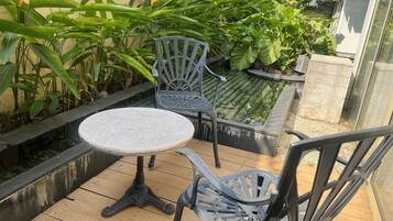 Una terraza de madera con una mesa redonda y dos sillas, rodeada de exuberante vegetación y una pequeña fuente de agua.