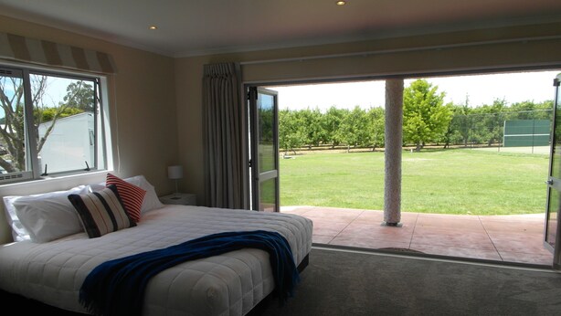 A bedroom with a bed, a nightstand, a lamp, and a view of a grassy area through a sliding glass door. - Cottages on St Andrews (Havelock North)