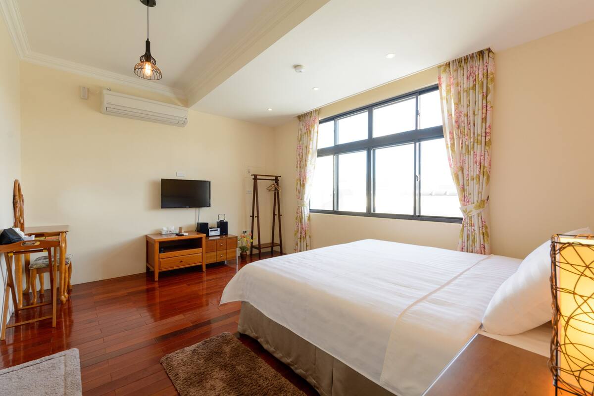 A bedroom with a large bed, a TV, a wooden dresser, and a desk.