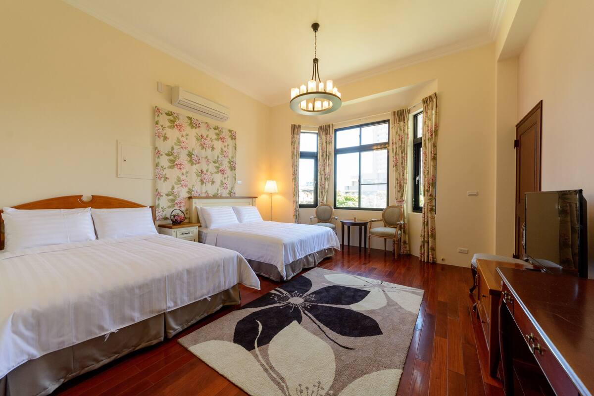 A hotel room with two beds, a chandelier, a small table with a lamp, a TV, and a floral wall decoration.