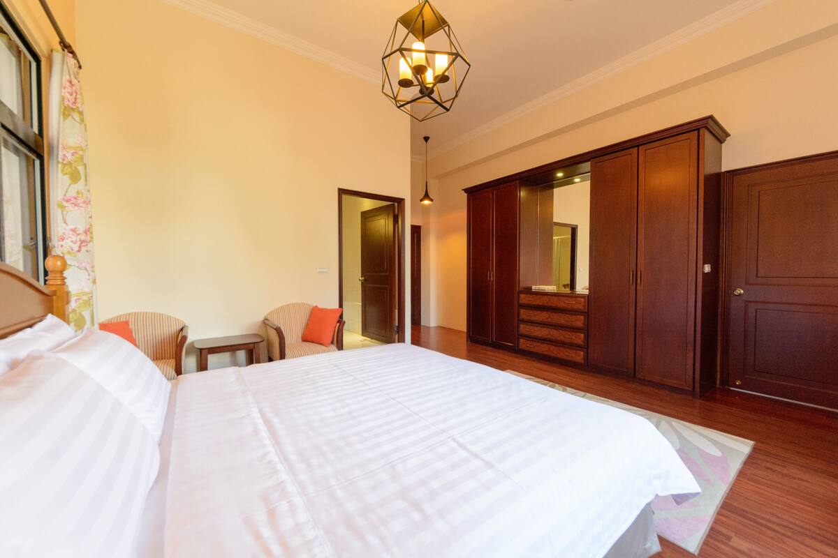 A hotel room with a large bed, a wooden wardrobe, a small table, and a chair.