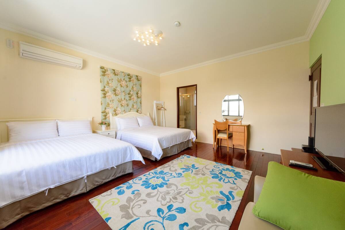 A hotel room with two beds, a desk, a chair, a TV, and a bathroom visible through a glass door.