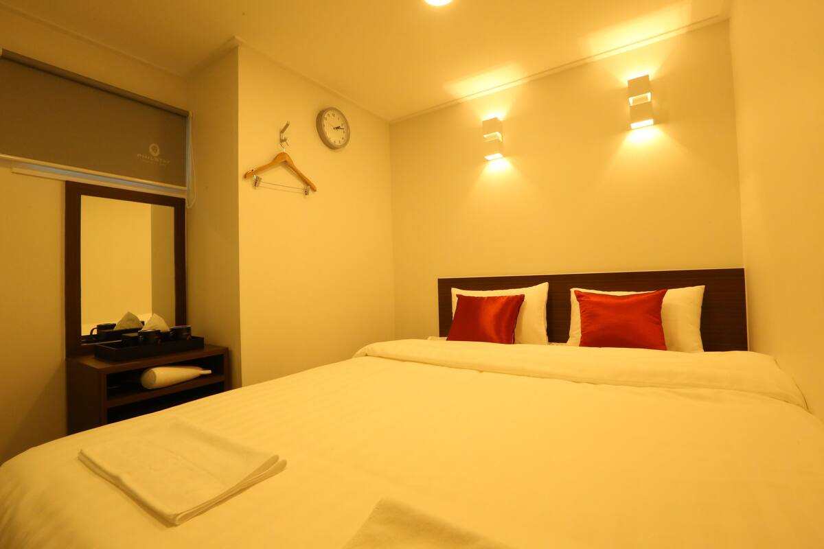 A hotel room with a bed, a bedside table, a mirror, a clock, and wall-mounted lights.