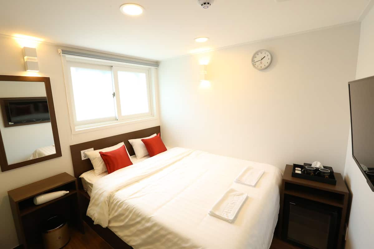A hotel room with a bed, a bedside table, a TV, a clock, and a window.